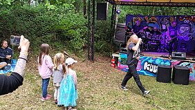 Vor einer Bühne singt ein Mann mit langen Haaren in ein Mikrofon. Drei Kinder schauen ihn an.