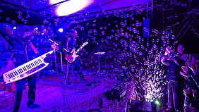 Zwei Musiker:innen rocken die Bühne. DAs Publikum jubelt. Im violetten Licht fliegen Seifenblasen.