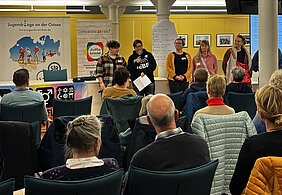 Fünf Menschen stehen vor dem nebeneinander sitzenden Publikum. Eine von ihnen hält ein Mikrofon in der Hand. Hinter ihnen befinden sich verschiedene Plakate.