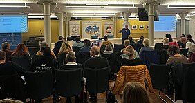 Menschen sitzen auf aufgereihten Stühlen nebeneinander. Vor ihnen steht ein Mann mit Mikrofon vor verschiedenen Plakaten.