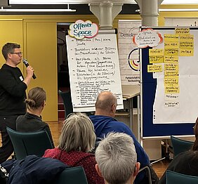 Menschen sitzen auf Am linken Bildrand steht ein Mann mit Mikrofon vor dem sitzenden Publikum. Neben ihm sind Plakate zum offenen Austauschraum aufgestellt.