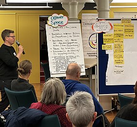 Menschen sitzen auf Am linken Bildrand steht ein Mann mit Mikrofon vor dem sitzenden Publikum. Neben ihm sind Plakate zum offenen Austauschraum aufgestellt.