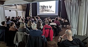 Das Publikum sitzt an Tischen und schaut auf eine Leinwand. Auf dieser wird der Dokumentarfilm "Stumpfe Sense - scharfer Stahl" gezeigt.