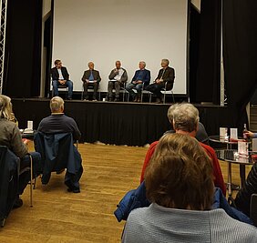 Auf einer Bühne sitzen fünf Männer. Sie führen eine Podiumsdiskussion. Das Publikum sitzt ihnen gegenüber an Tischen. 