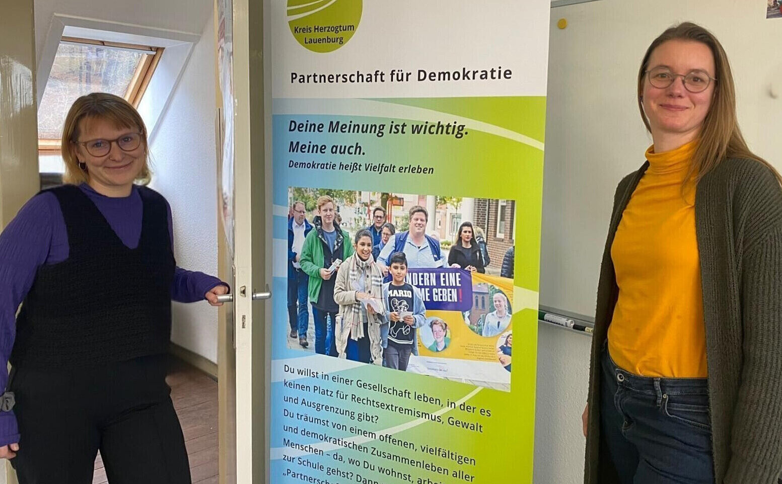 Maren Schumann hält die Klinke einer geöffneten Tür in der Hand und lächelt. Ein Roll-Up beschreibt die Partnerschaft für Demokratie. Rechts im bild steht Rike Nagel.